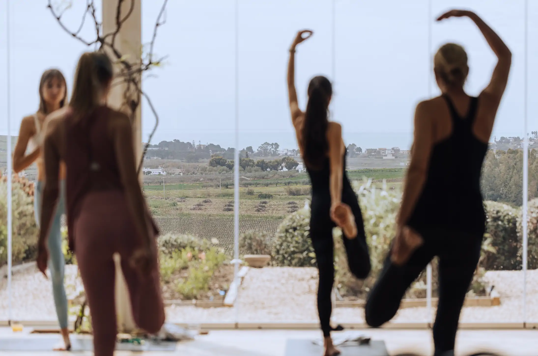 Spring yoga retreat. Participants practicing yoga in a serene, light-filled space at La Segreta, with panoramic views of lush vineyards and the Mediterranean Sea in the distance. This image promotes the Spring Retreat for the Senses by Alexandra Meffert, hosted at the Planeta Family Estate, combining mindful movement, relaxation, and a deep connection to nature in an idyllic Sicilian setting.